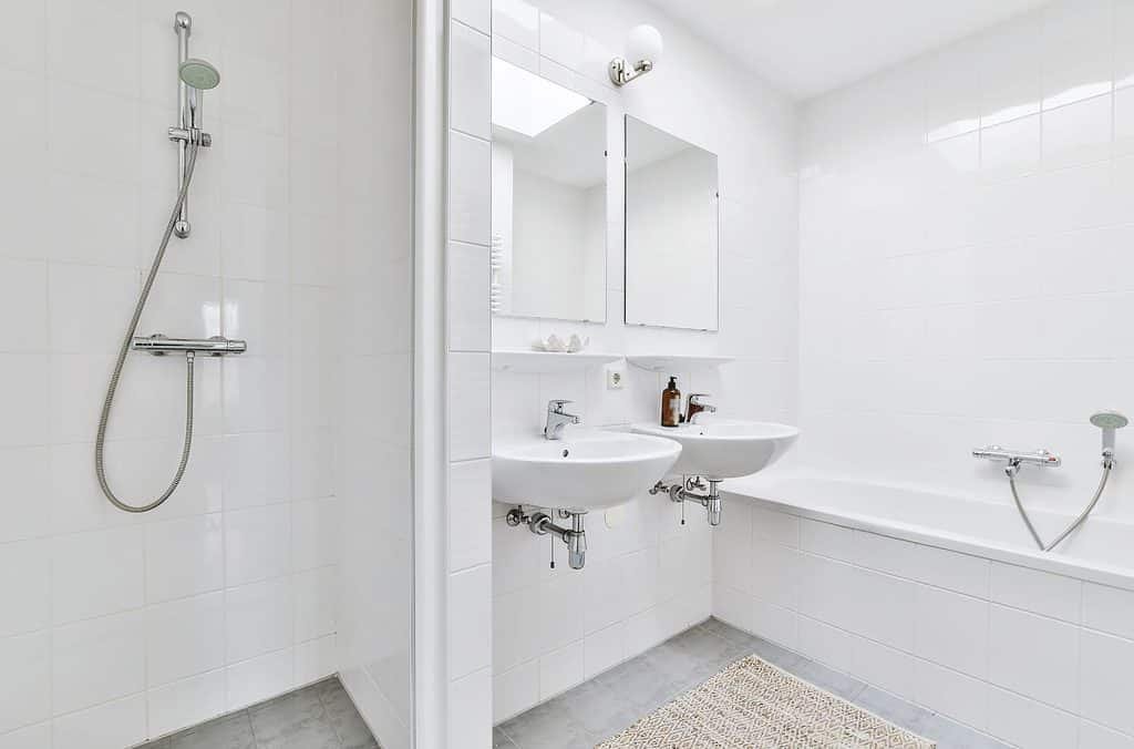 Wet room with white tiles - a shower on the left, two sinks and a bath with a shower