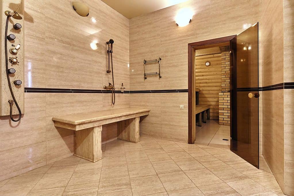 A large marble wet room with a shower on the left and a stone seat in the centre and a doorway through to what appears to be a steam room